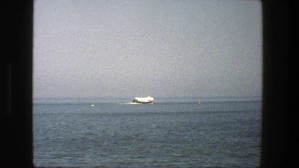 A distant white boat moves across calm Danish water in a 1975 8mm home-movie frame with muted color and soft focus.