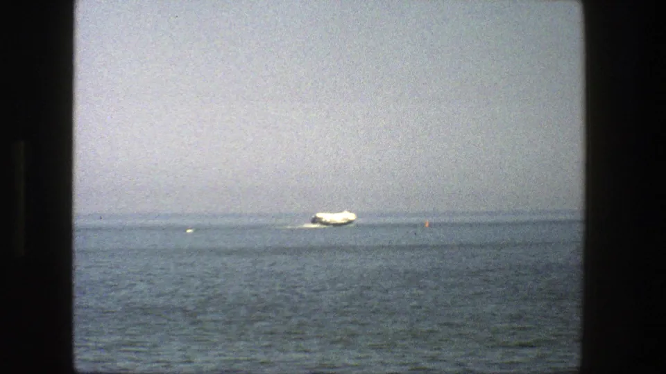 A white boat sits near the horizon on calm water in Denmark, framed by dark 8mm film edges and a pale sky.