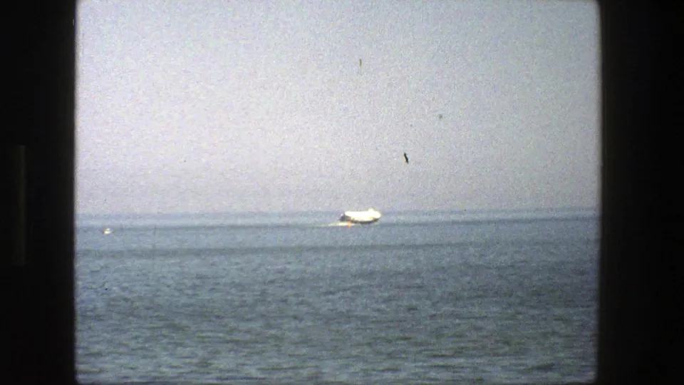 1975 Denmark home-movie frame with a distant white boat on calm water, a hazy horizon, and a dark window edge in the foreground.