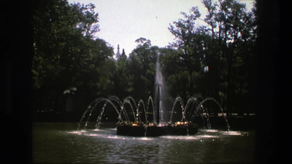 1975 Denmark 8mm frame showing a park fountain spraying arcing jets over a pond, with dense green trees behind it.