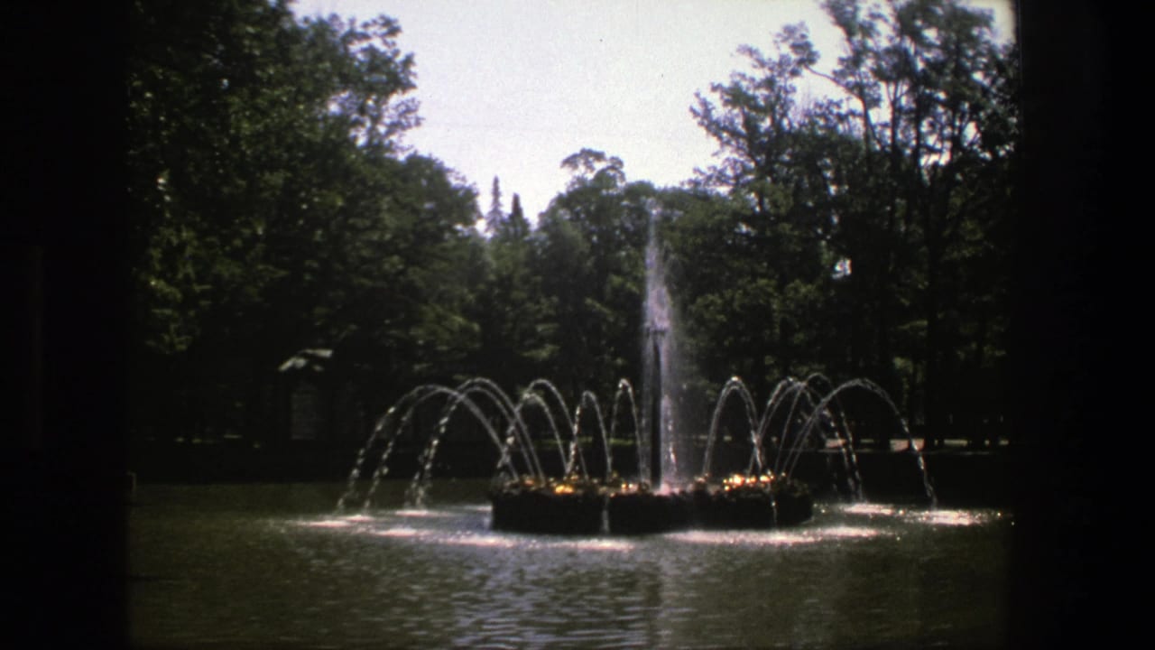 1975 Denmark 8mm frame showing a tree-lined park fountain with arcing water jets and a central spray, seen through dark film or window edges.