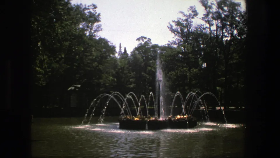 1975 Denmark 8mm frame showing a tree-lined park fountain with arcing water jets and a central spray, seen through dark film or window edges.