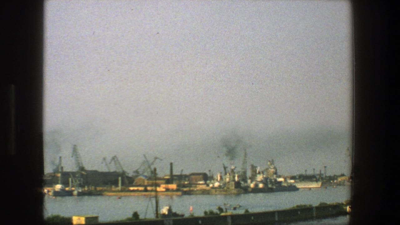 Distant harbor with cranes, dock structures, ships, water, and pale sky.