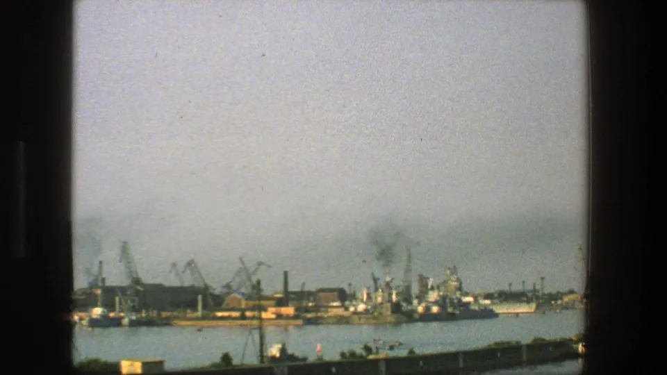 1975 Denmark 8mm frame showing a distant waterfront with cranes, ships, industrial dock structures, and haze over the harbor.
