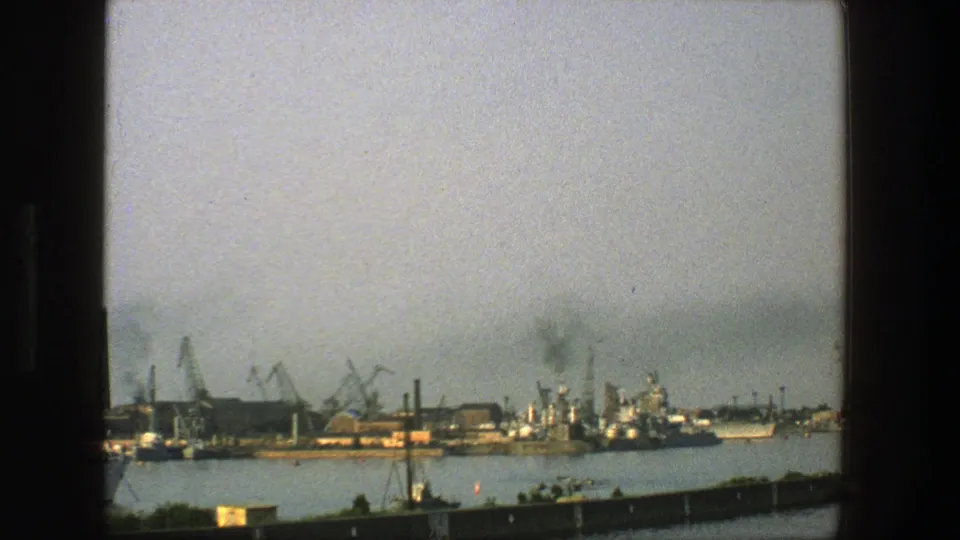 1975 Denmark 8mm frame showing an industrial waterfront with cranes, moored ships, dock buildings, and a dark haze above the harbor.