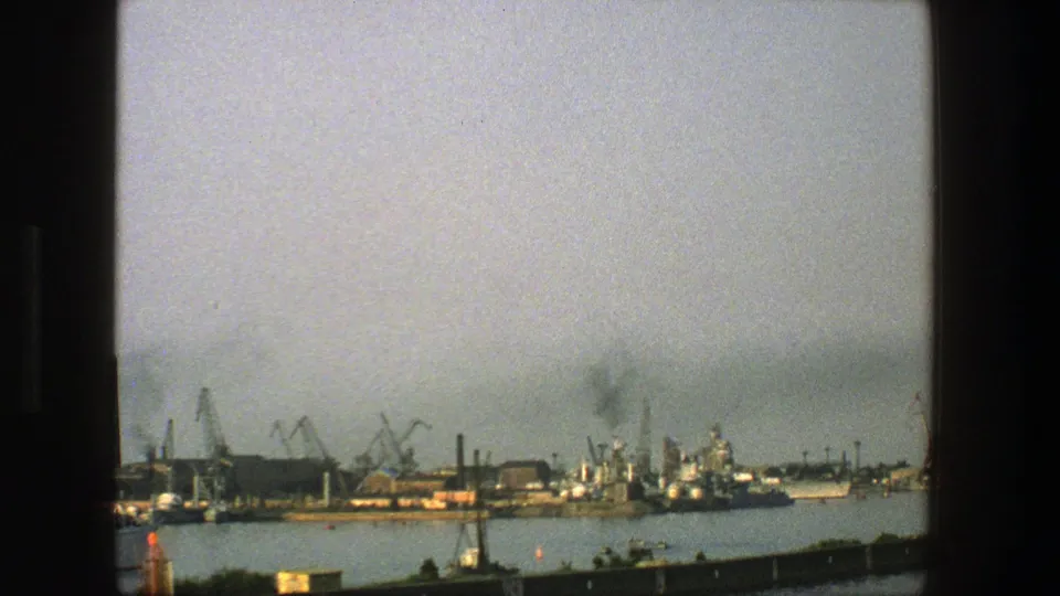 1975 Denmark 8mm frame showing a broad harbor with cranes, vessels, industrial docks, foreground shoreline, and smoke or haze in the sky.