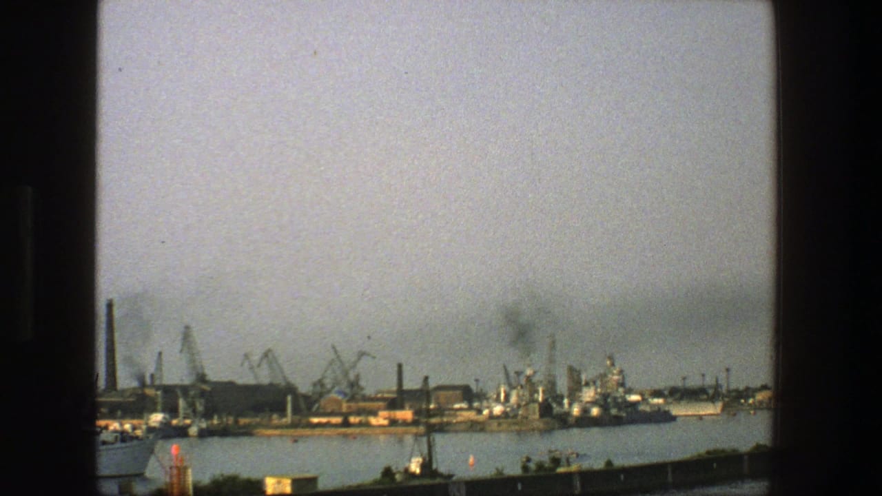 Industrial harbor across calm water with cranes, smokestacks, ships, and haze.