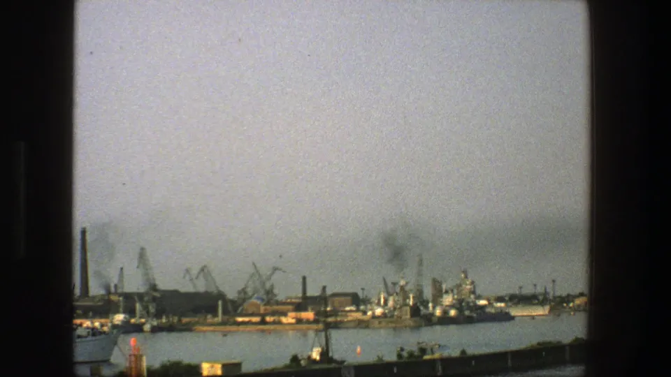 1975 Denmark 8mm frame showing an industrial harbor with cranes, smokestacks, moored vessels, and calm water.