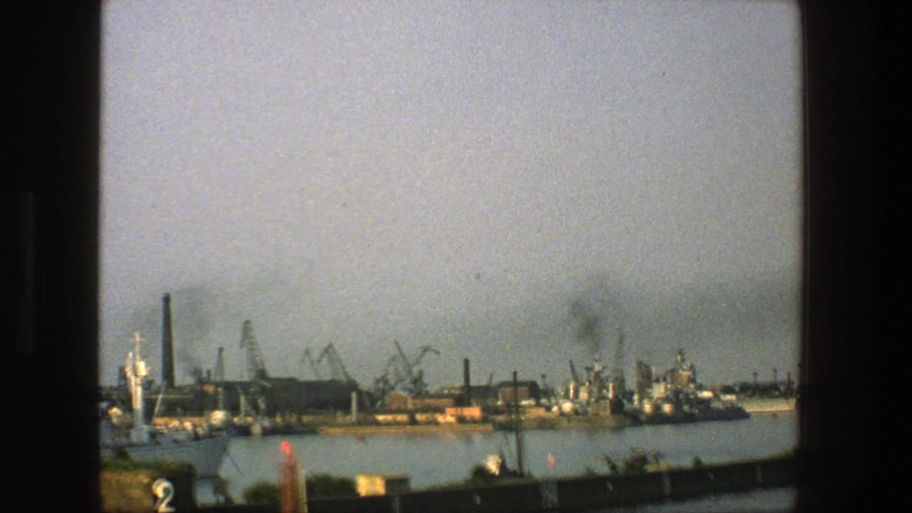 1975 Denmark 8mm frame with a broad industrial waterfront, dock cranes, smokestacks, and vessels along calm water.