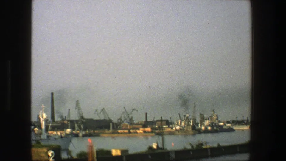 1975 Denmark 8mm frame with a broad industrial waterfront, dock cranes, smokestacks, and vessels along calm water.