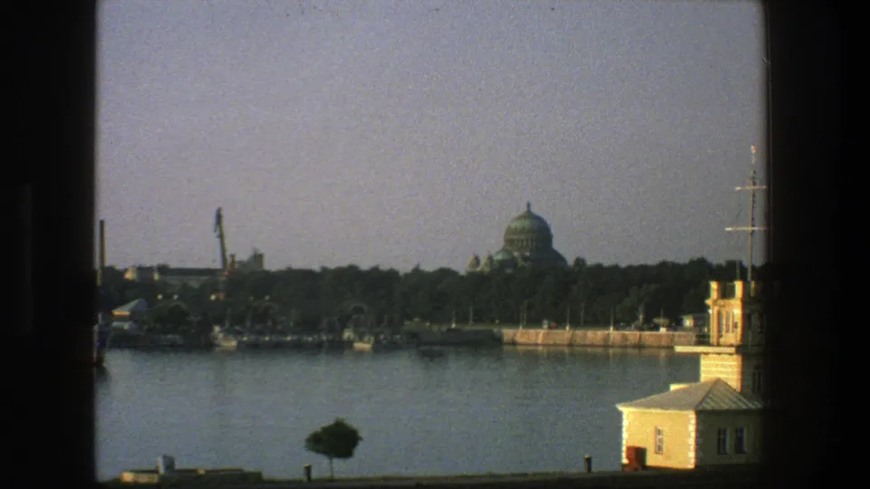 1975 Denmark 8mm frame showing calm water, tree-lined shore, moored boats, a small waterfront building, and a distant domed structure.