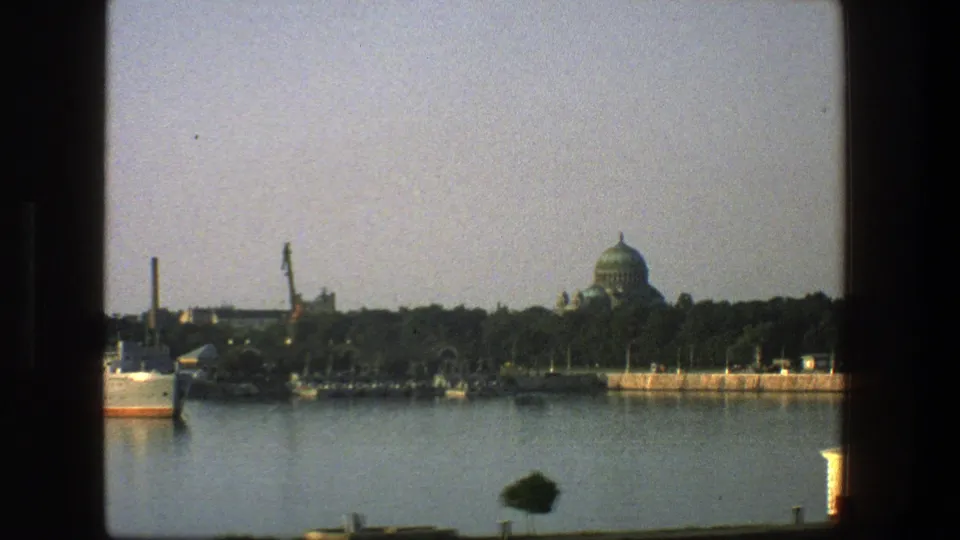1975 Denmark 8mm frame of a quiet waterfront with trees, boats, industrial stacks, and a distant domed building across calm water.