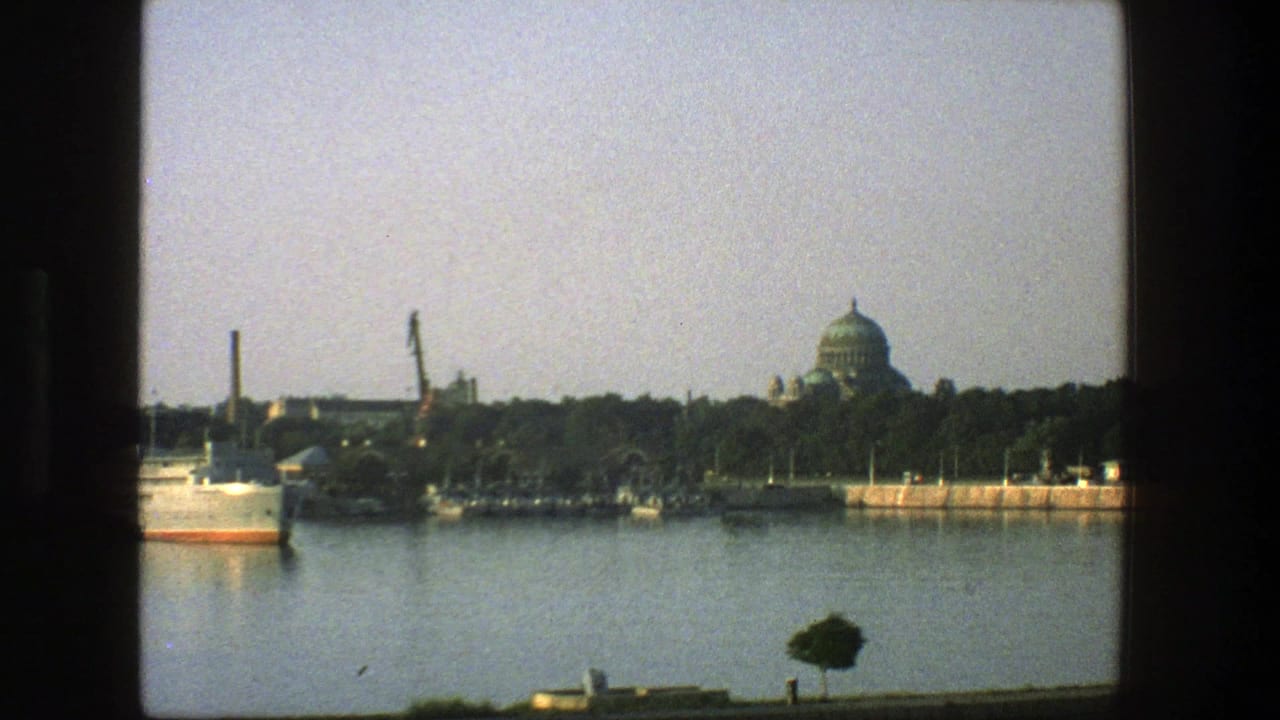 Large ship on calm water with cranes, trees, smokestack, and green-domed building beyond.