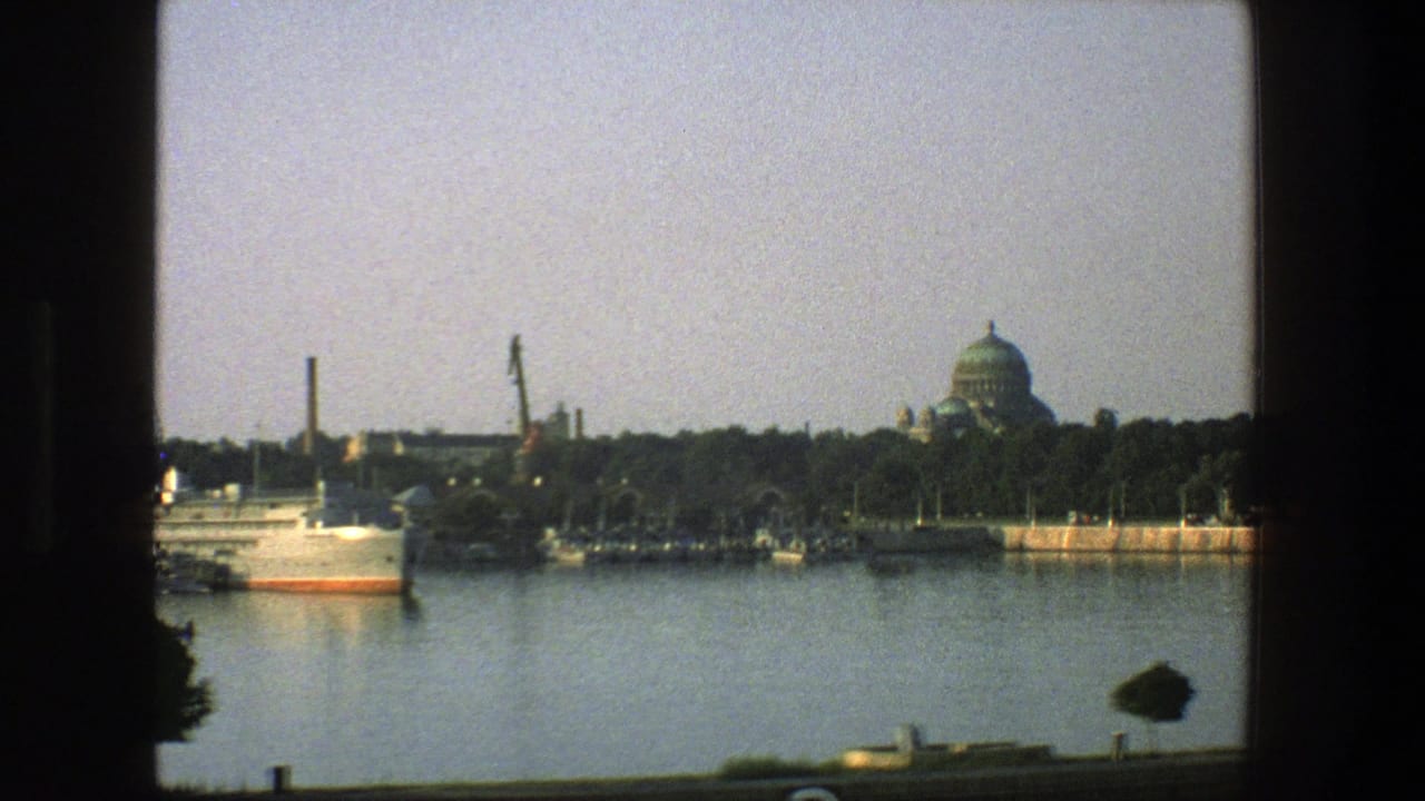 White ship near a harbor shoreline with cranes, trees, smokestack, and domed building.