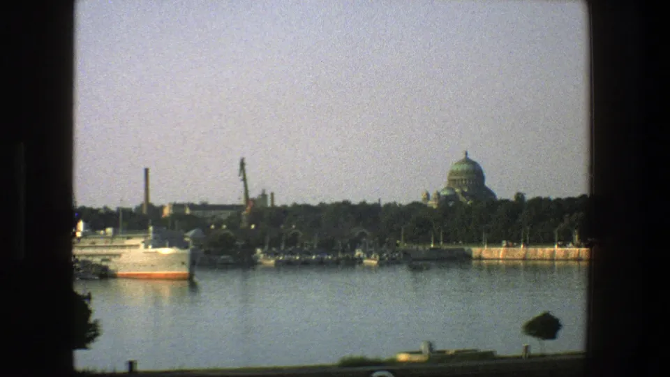 1975 Denmark home-movie frame of a white ship moving along a port waterfront with cranes and a green-domed building behind trees.