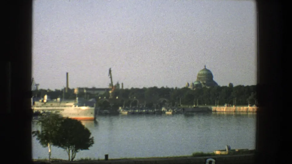 1975 Denmark 8mm frame with the ship partly visible at left, calm water in front, and cranes and a green-domed building across the shore.