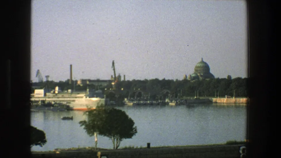 1975 Denmark frame showing a harbor view after the larger ship has moved on, with cranes, a small boat, trees, and a green-domed building.