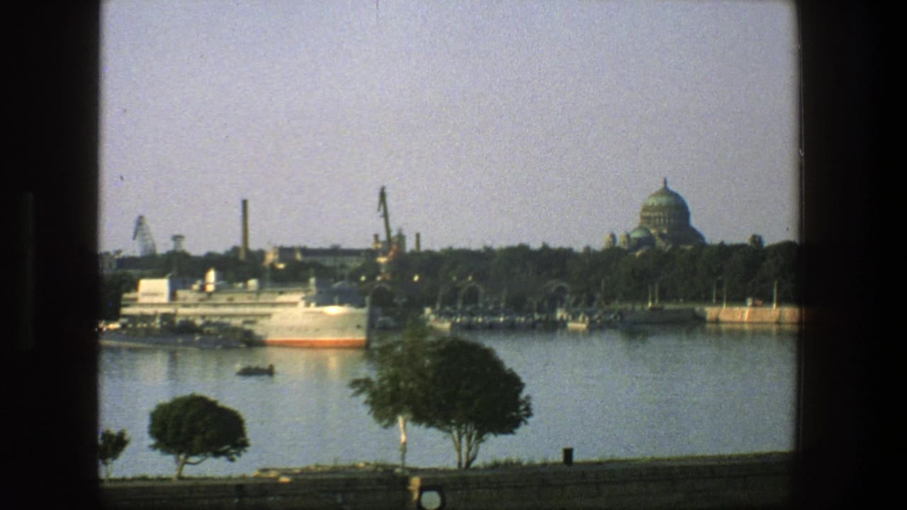 Docked vessel in a calm harbor channel with cranes, trees, and a distant domed building.
