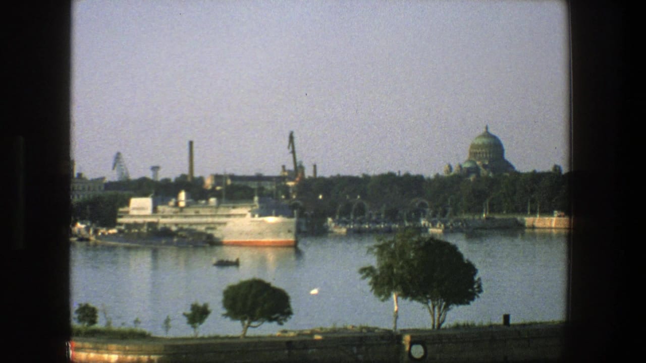 Harbor view with a docked ship, small boat, cranes, trees, and a domed building in the distance.