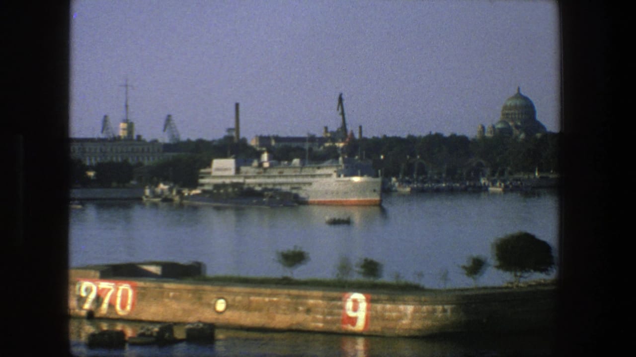 1975 Denmark harbor seen through a vessel window, with a docked ship, cranes, water, and a domed waterfront building beyond.