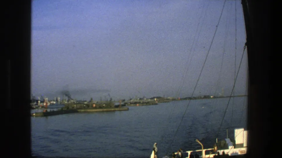 Shipboard view across a Denmark harbor, with distant vessels and cranes, rigging in the foreground, and a small person on deck.