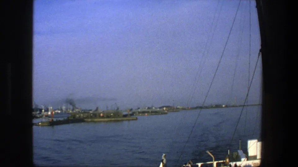 1975 Denmark harbor scene from aboard a vessel, with water, distant docked ships, cranes, and rigging crossing the frame.