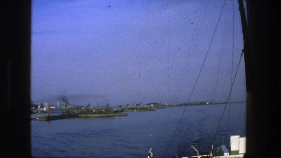 A Denmark harbor viewed from a moving vessel, showing distant ships and cranes across blue water with rigging and deck equipment nearby.