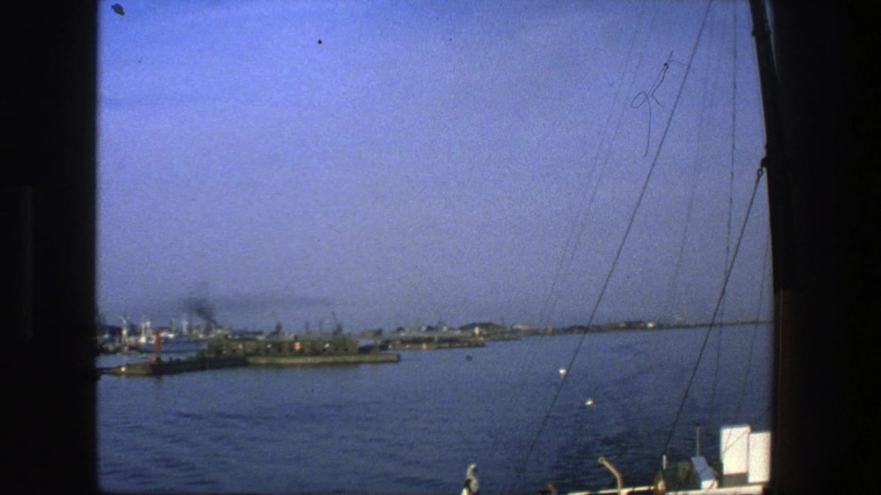 1975 Denmark 8mm frame looking from a boat across calm blue water toward low harbor buildings and smokestacks.