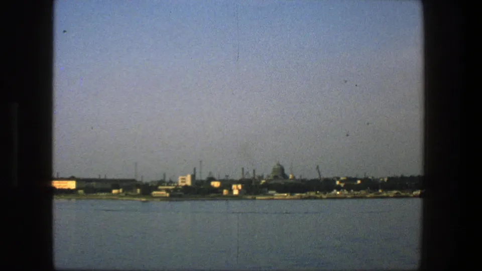 1975 Denmark home-movie frame of a distant waterfront skyline across water, with industrial silhouettes and a dome-like structure.