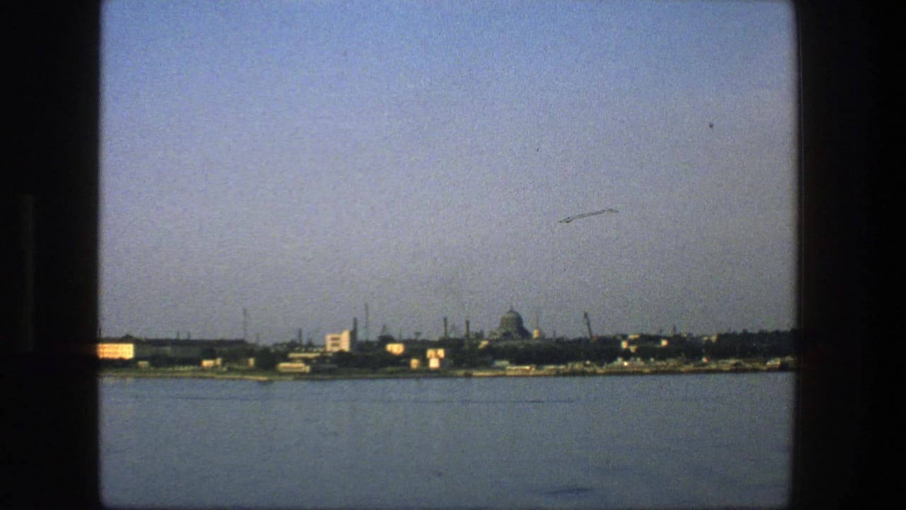 1975 Denmark 8mm view across water toward a low harbor skyline, with a distant domed form and industrial waterfront details.