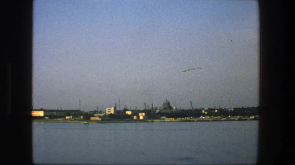 1975 Denmark 8mm view across water toward a low harbor skyline, with a distant domed form and industrial waterfront details.