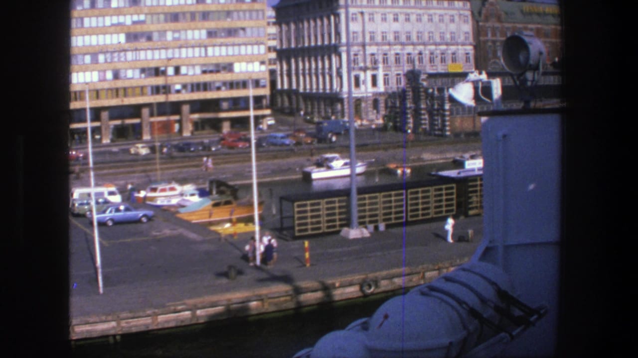 Waterfront quay with small boats, parked cars, pedestrians and multi-story buildings.