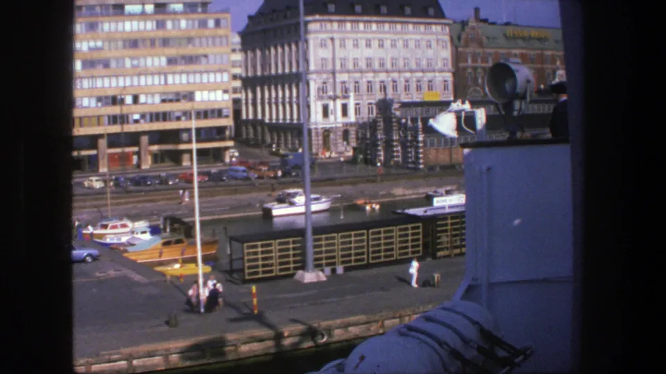 1975 Denmark waterfront frame showing small boats along the quay, pedestrians near the water and dense urban buildings in the background.