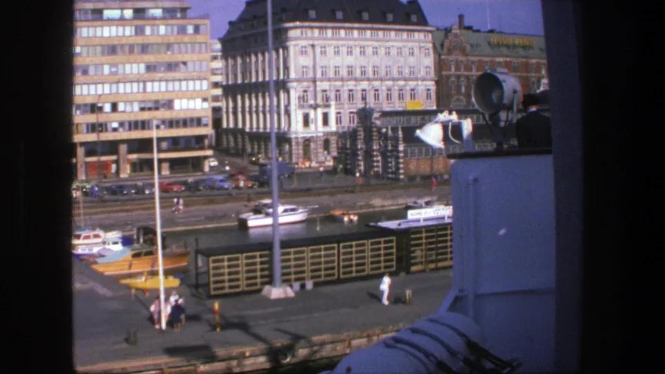 1975 Denmark harbor scene with boats moored along the quay, pedestrians on the pier and classic waterfront buildings beyond.