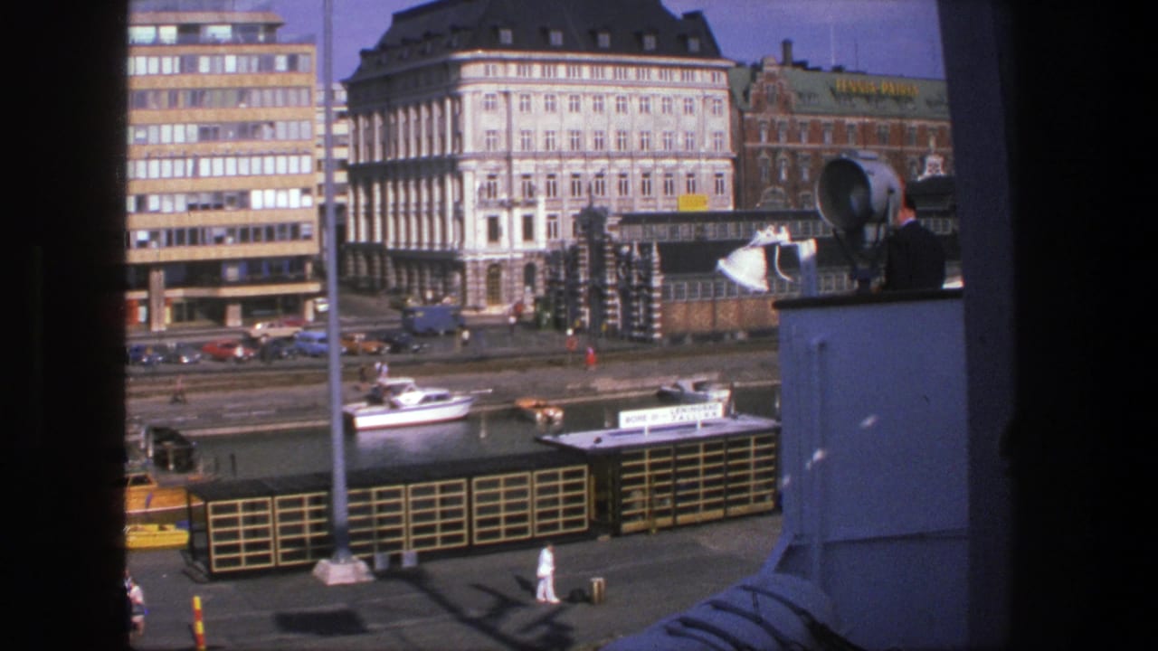 1975 Denmark 8mm view over an urban waterfront with tall buildings, a small boat, quay railings, and pedestrians.