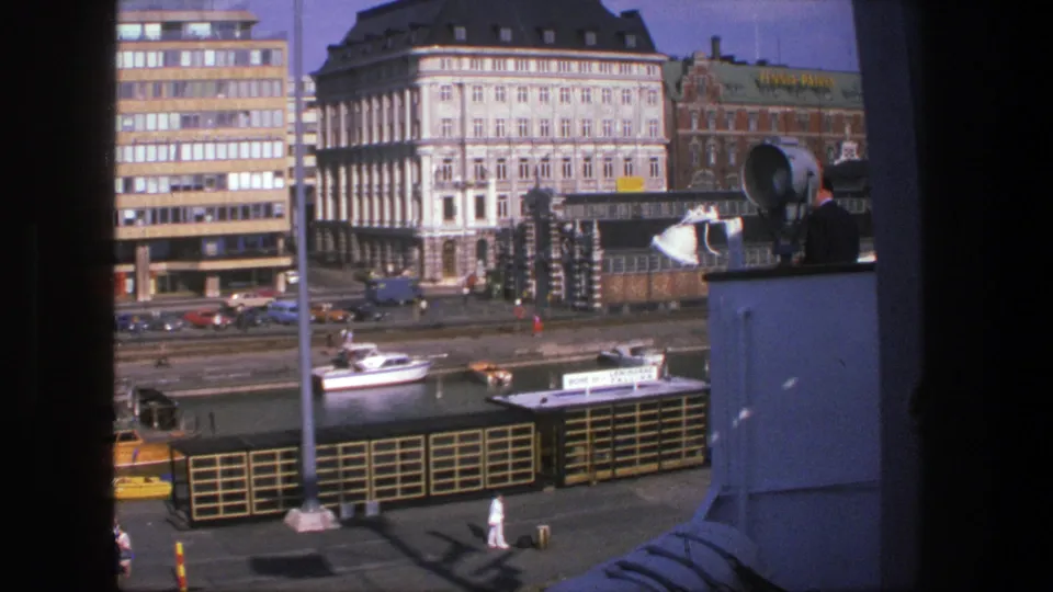 1975 Denmark 8mm view over an urban waterfront with tall buildings, a small boat, quay railings, and pedestrians.
