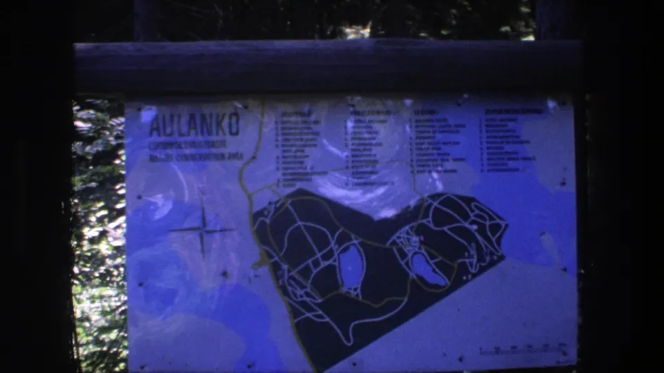 The camera holds on a posted Aulanko conservation-area trail map, with rail and water reflections around the sign.