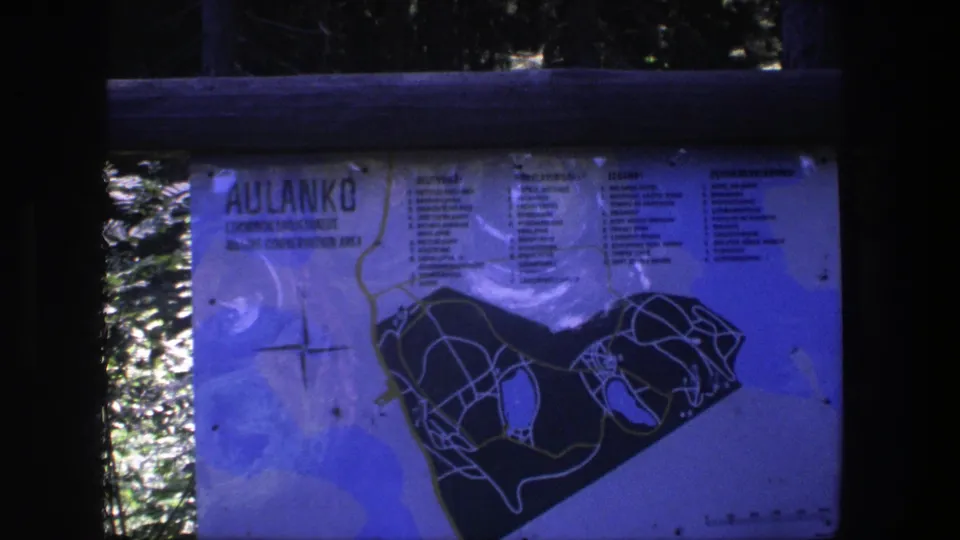 1975 8mm frame of an outdoor Aulanko park map sign in Finland, seen through wooden rails with trees behind it.
