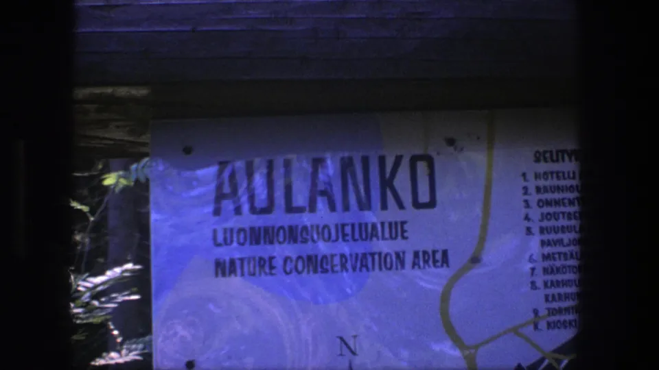 Blue-cast 8mm frame of the Aulanko Nature Conservation Area sign, including Finnish wording, English label, and a cropped visitor list.