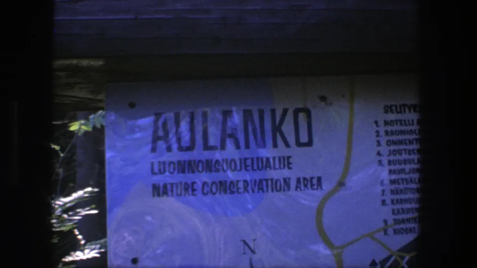 Final still from the 8mm clip showing the Aulanko Nature Conservation Area sign, dark surrounding frame, and partial foliage at the edge.