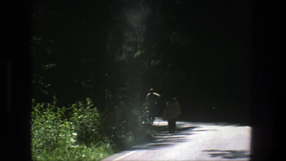 Two people walk away along a shaded Danish roadside in 1975 8mm home-movie footage, with bright sun on the pavement.