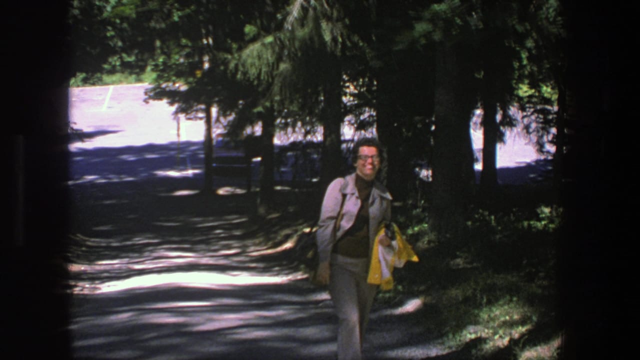 Woman in gray suit walks up a shaded park path carrying a yellow jacket.