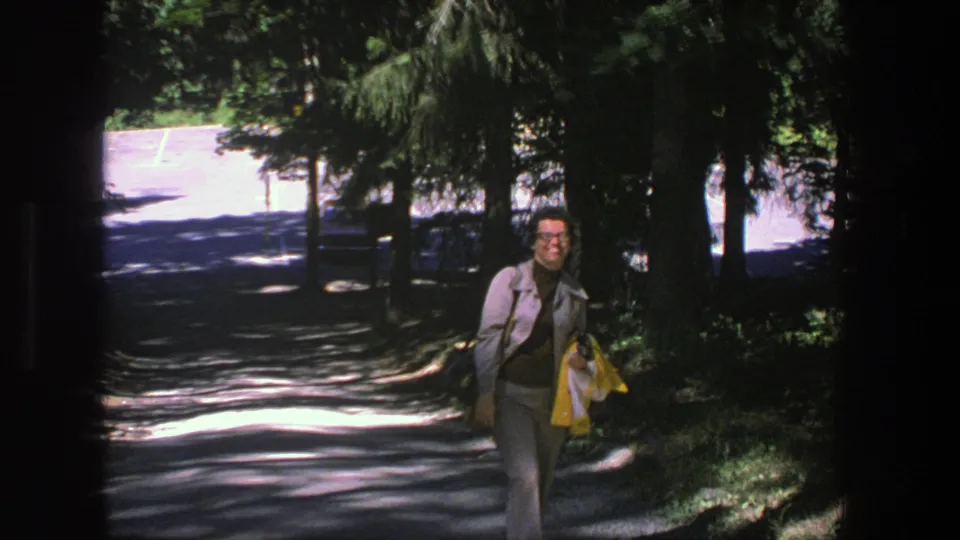 In Denmark 1975, a woman in glasses and a gray suit walks along a tree-lined path carrying a yellow jacket.