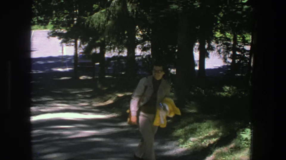 The woman in a gray suit continues along the Denmark park path, holding a yellow jacket as trees cast deep shade.