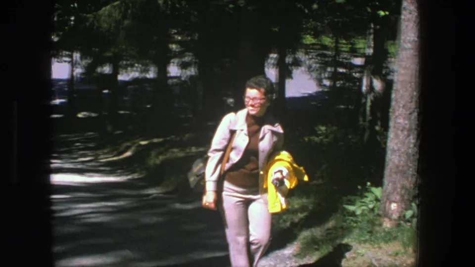 A smiling woman in glasses walks closer to the camera in Denmark, wearing a gray suit and carrying a bright yellow jacket.