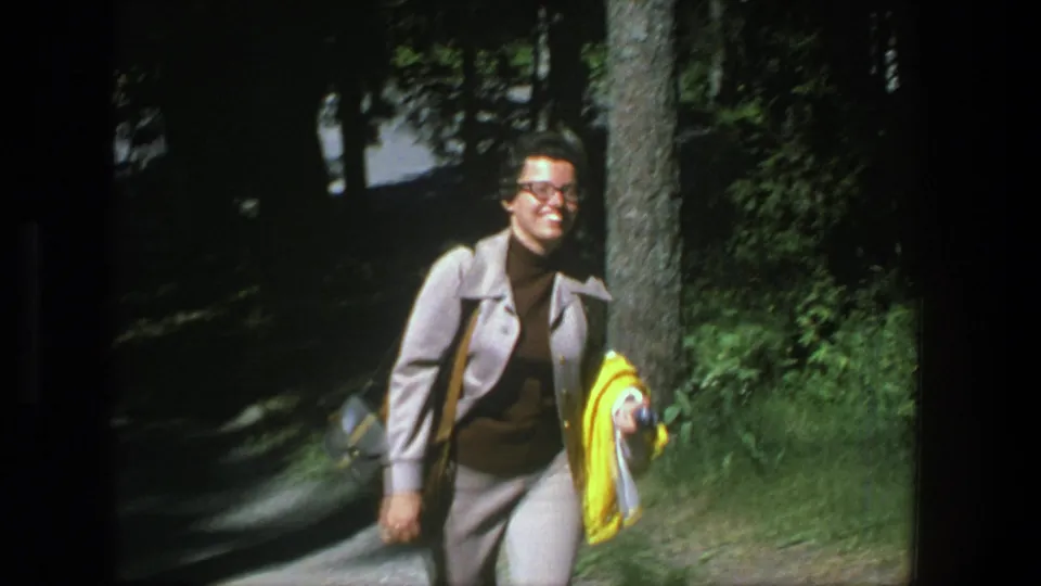 Near the end of the Denmark 1975 clip, the woman smiles close to the camera beside trees and a shaded green verge.