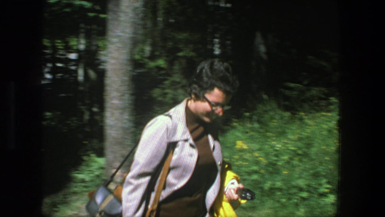 Woman with glasses walking beside trees while holding a yellow jacket.