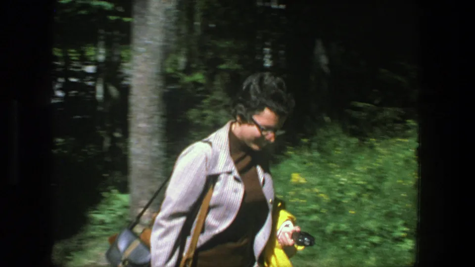 In Denmark, 1975, a woman in glasses and layered clothing walks through a sunlit forest holding a yellow jacket.