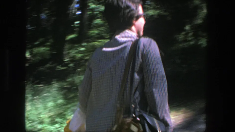 The woman is seen from behind on a forest path, carrying a shoulder bag as the 8mm camera tracks her movement.