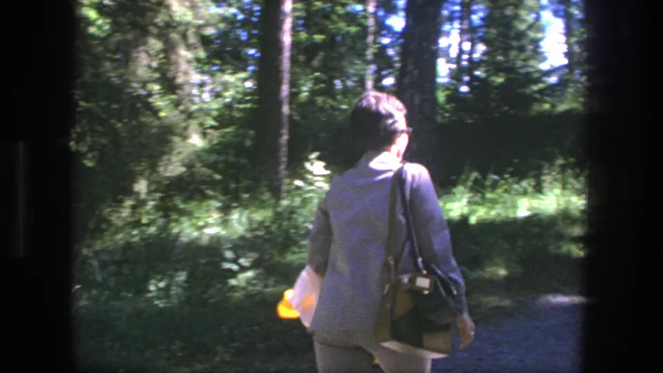 In a sun-dappled Danish woodland, the woman walks away with a shoulder bag and yellow jacket near a narrow path.